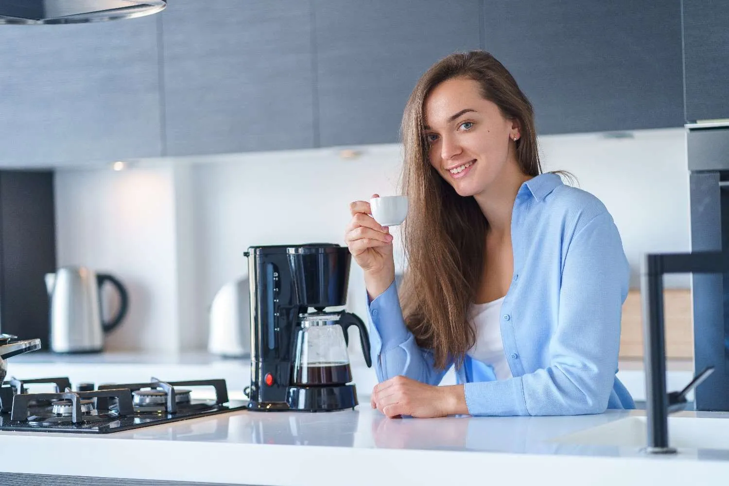 Smiling woman holding a cup of coffee beside the coffee maker in her kitchen